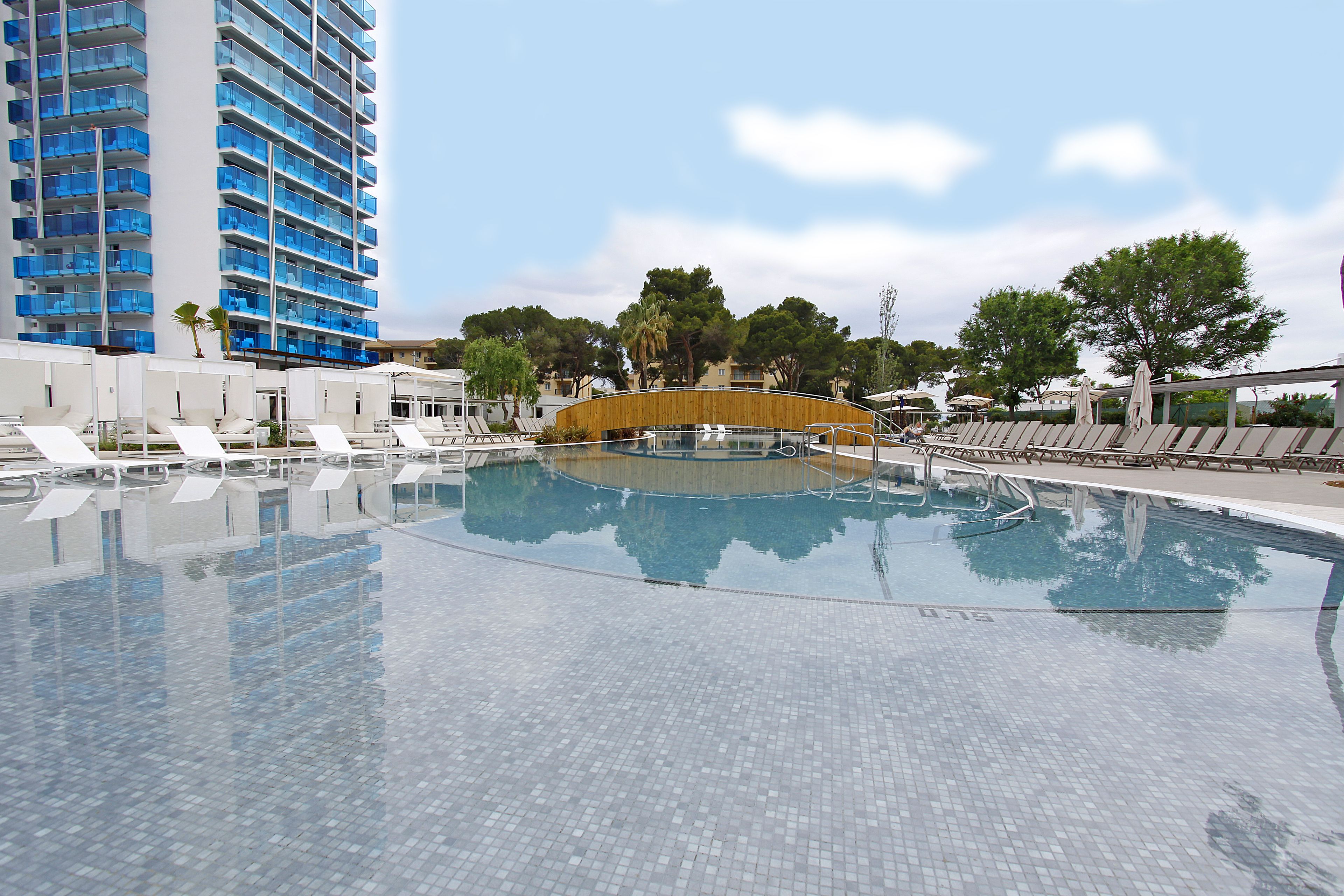 Una piscina con sillones de descanso, un puente de madera y un edificio alto al fondo.