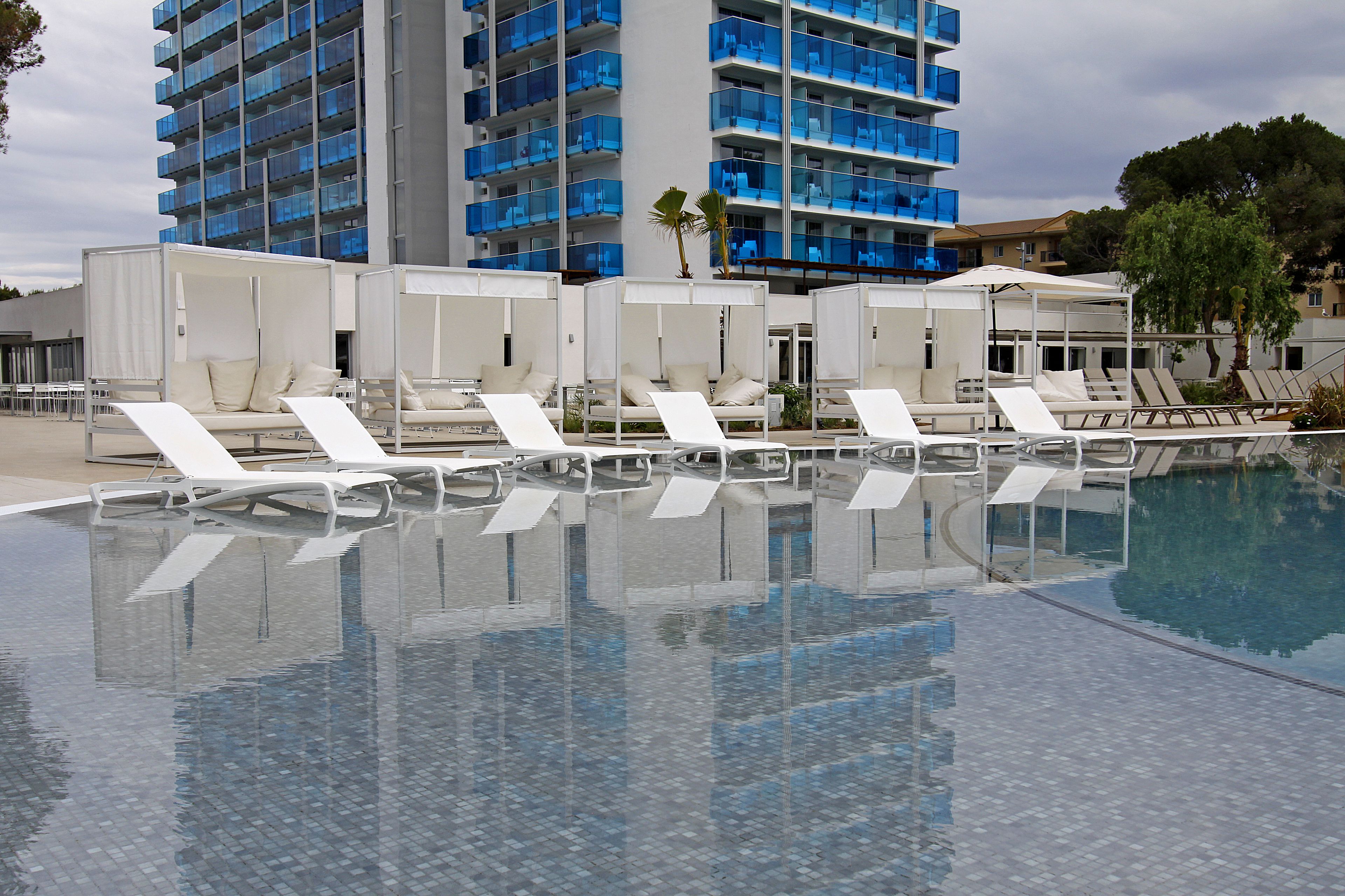 A pool area with white sun loungers and a large building with blue balconies in the background.