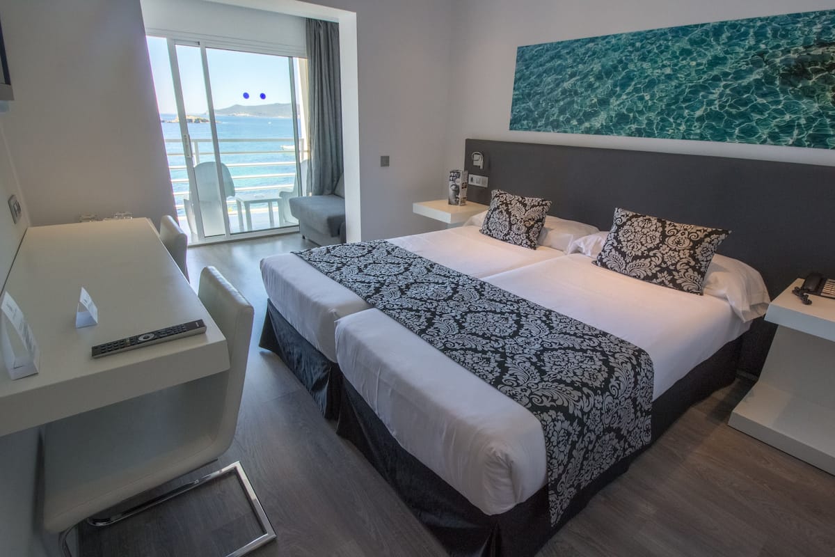 Une chambre d’hôtel avec un lit, un bureau et une chaise. La fenêtre offre une vue sur la mer.