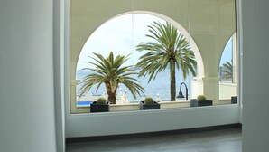 Een kamer met een groot, rondboogvormig raam dat uitkijkt op een balkon met palmbomen en uitzicht op zee.