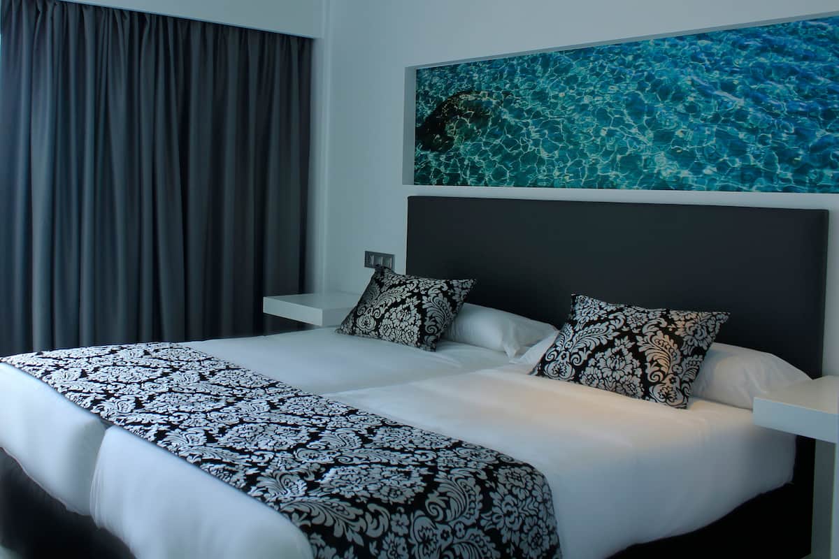 Une chambre d’hôtel moderne avec un grand lit, des rideaux foncés, une œuvre d’art accrochée au mur et une table de chevet blanche.