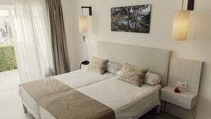 A double bed with a beige bedspread and two patterned pillows, a nightstand with a red rose, and a painting of a landscape on the wall. - Hotel d´Or (Santanyi)