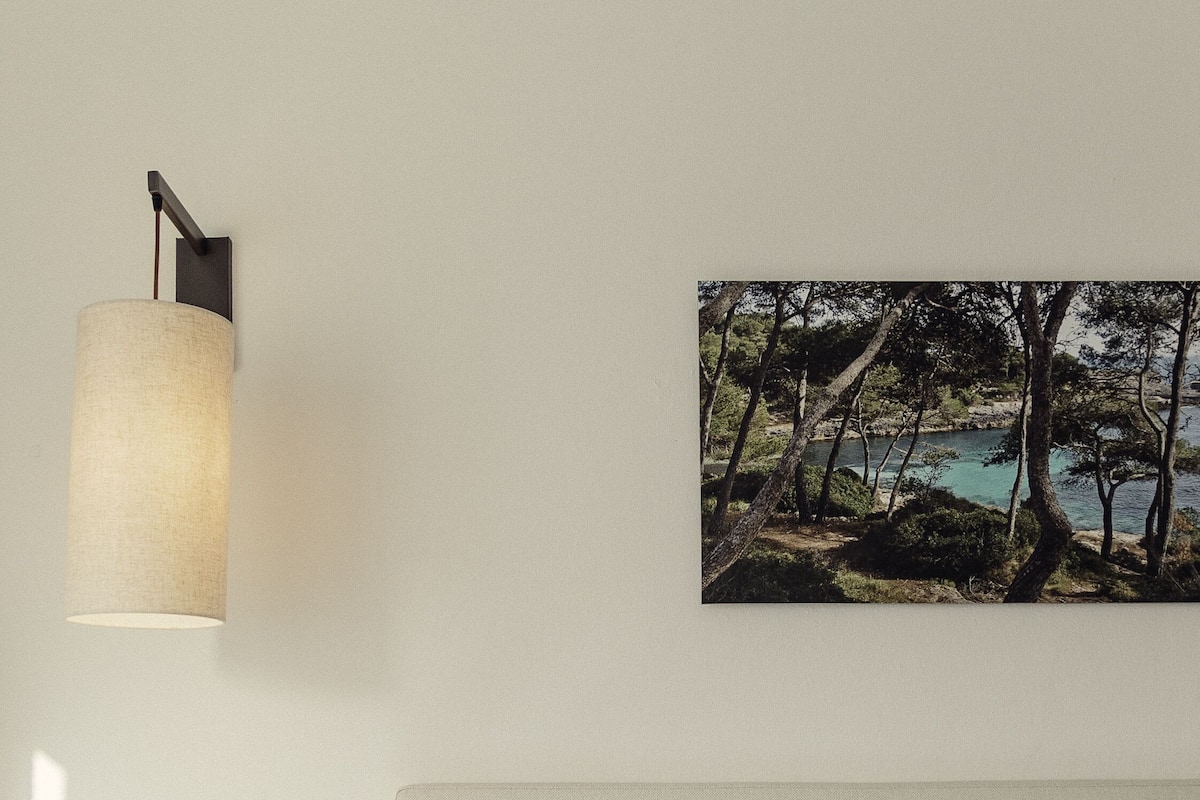 A bedroom with a bed, a nightstand with a phone, a lamp, and a framed picture of a landscape.