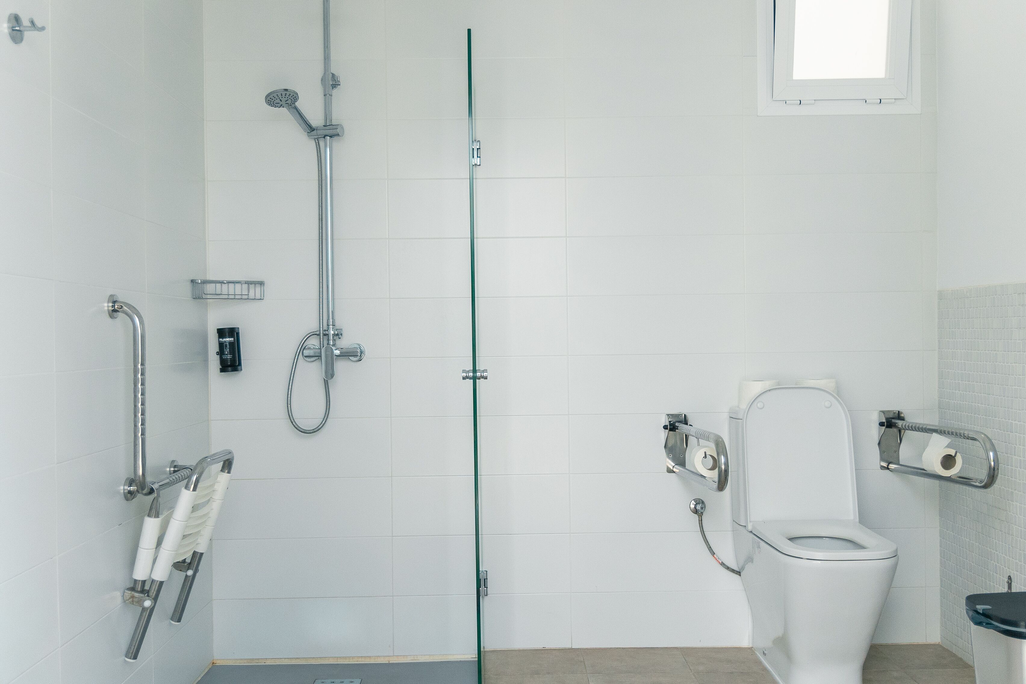A modern bathroom with a glass shower enclosure, a white toilet with grab bars, and a small window.
