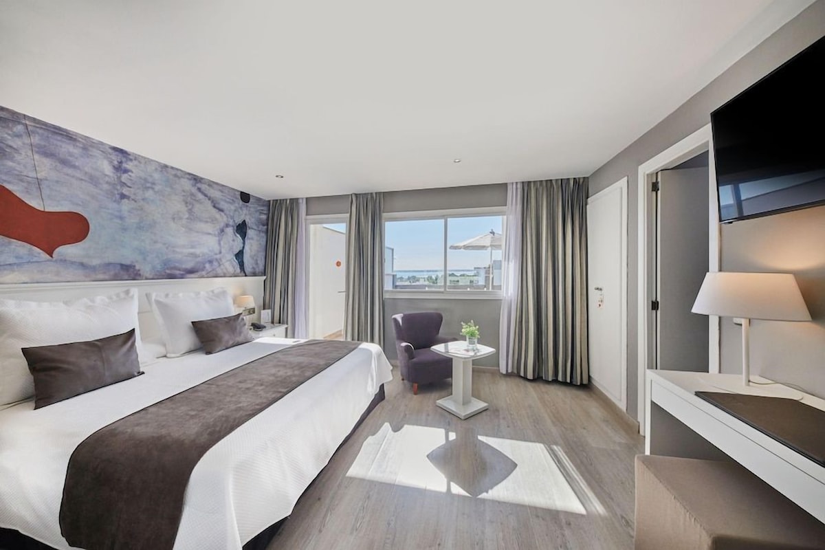 A hotel room with a bed, a chair, a small table, a TV, and a window with curtains.