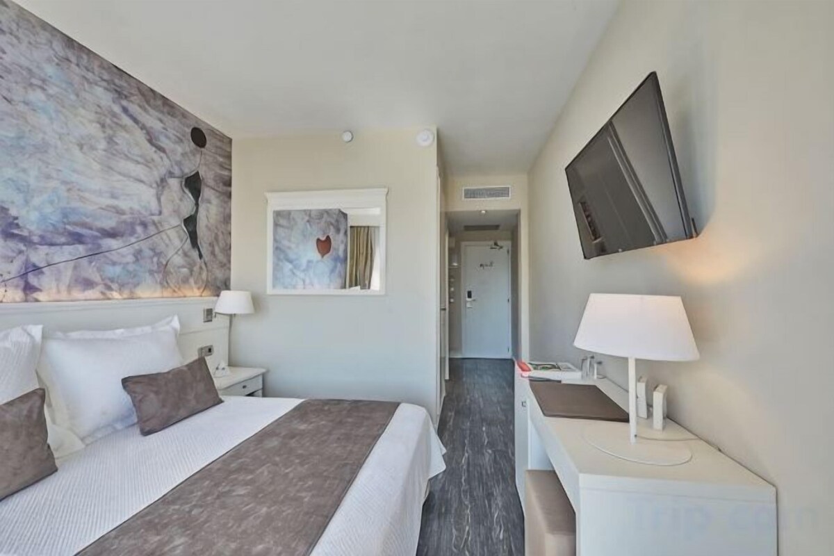 Habitación de hotel con cama, mesitas de noche, escritorio, televisor y obras de arte en las paredes.