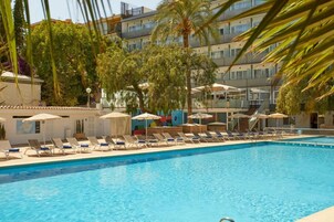 Una piscina con tumbonas y sombrillas, rodeada de árboles y un edificio de varios pisos.