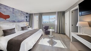 Una habitación de hotel moderna con una cama grande, una mesita con una lámpara, una silla y un televisor montado en la pared.