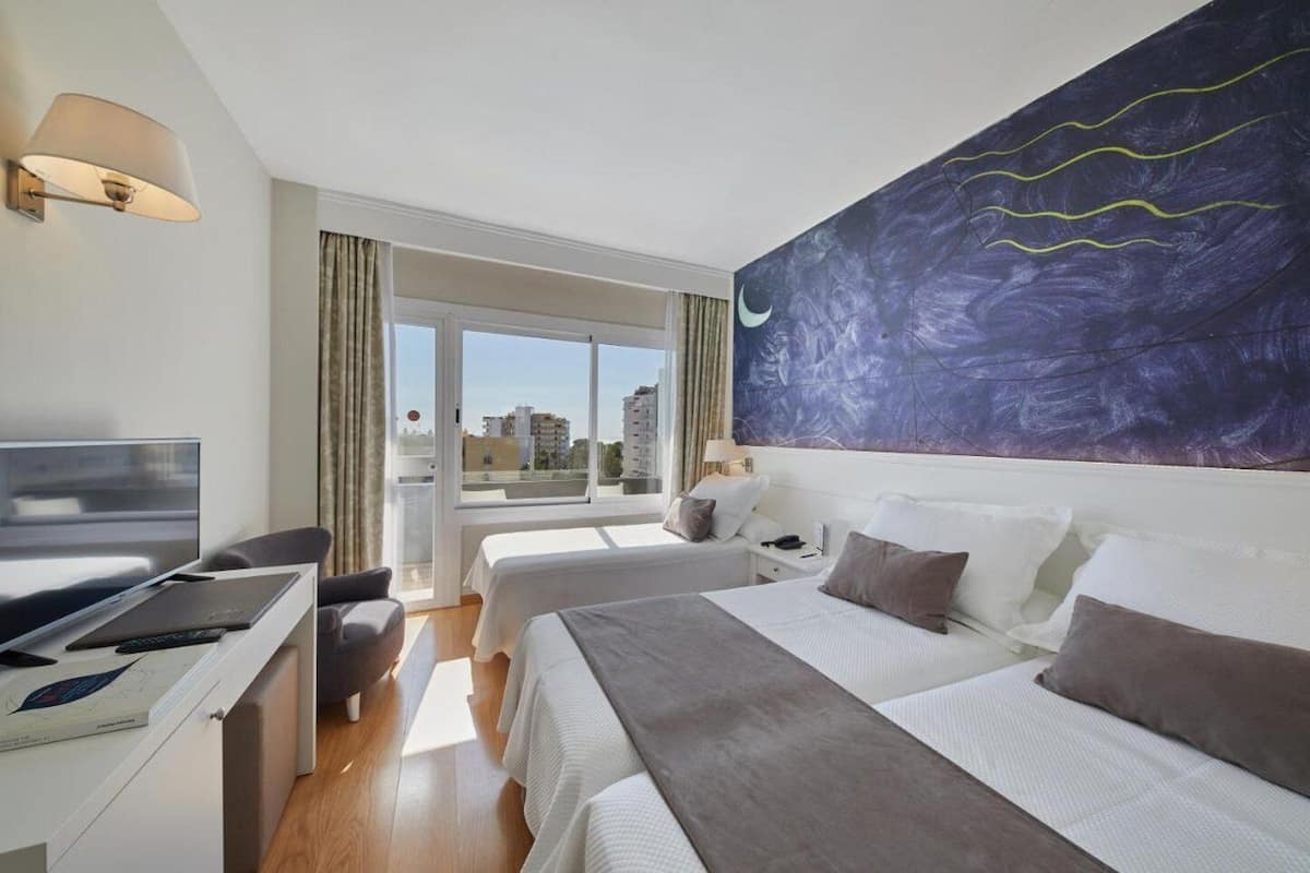 Habitación de hotel con dos camas, un escritorio con una laptop, un televisor y un ventanal con vistas a la ciudad.