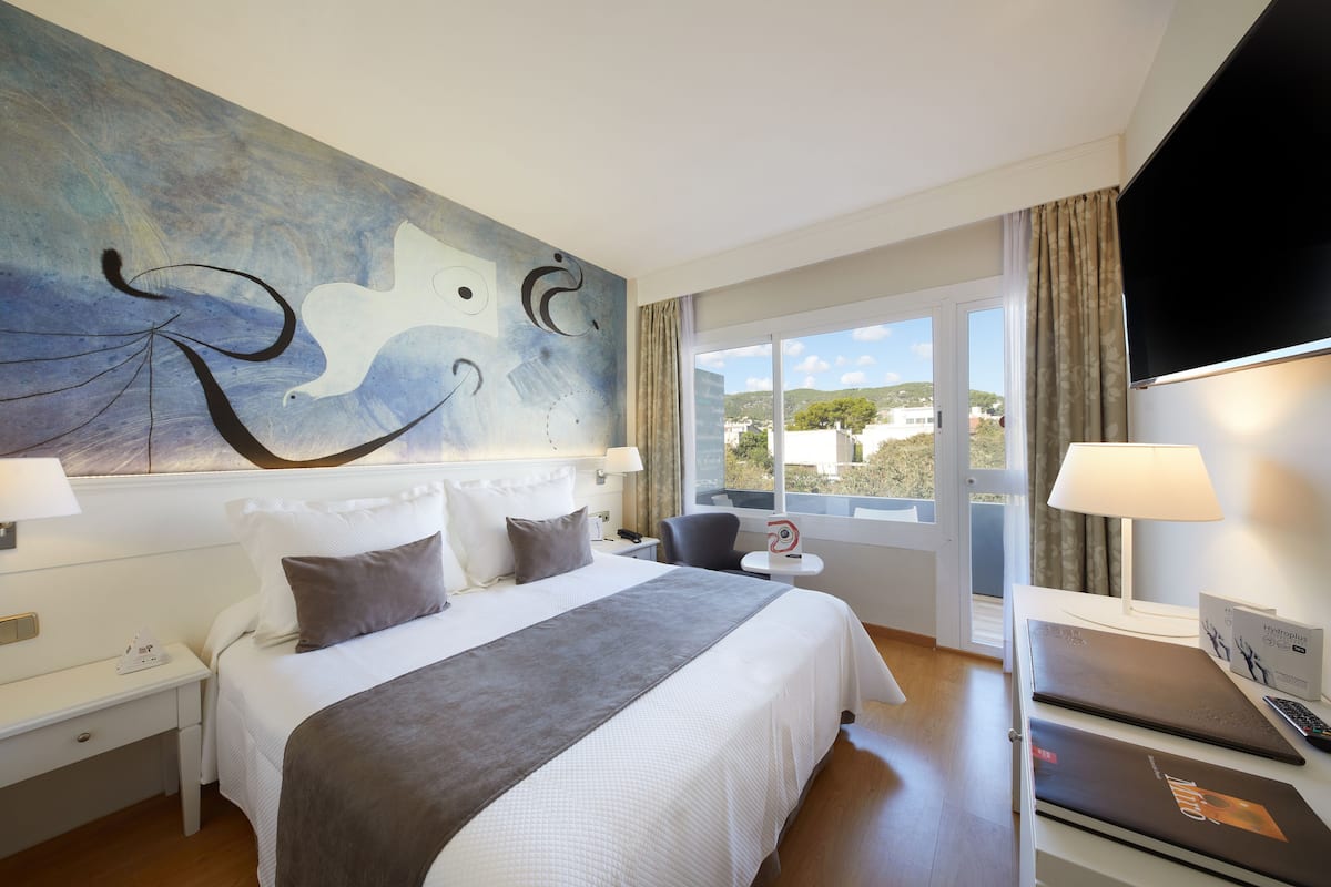 Habitación de hotel con una cama grande, un escritorio con una computadora portátil, un cuadro en la pared y una ventana con vistas a edificios.