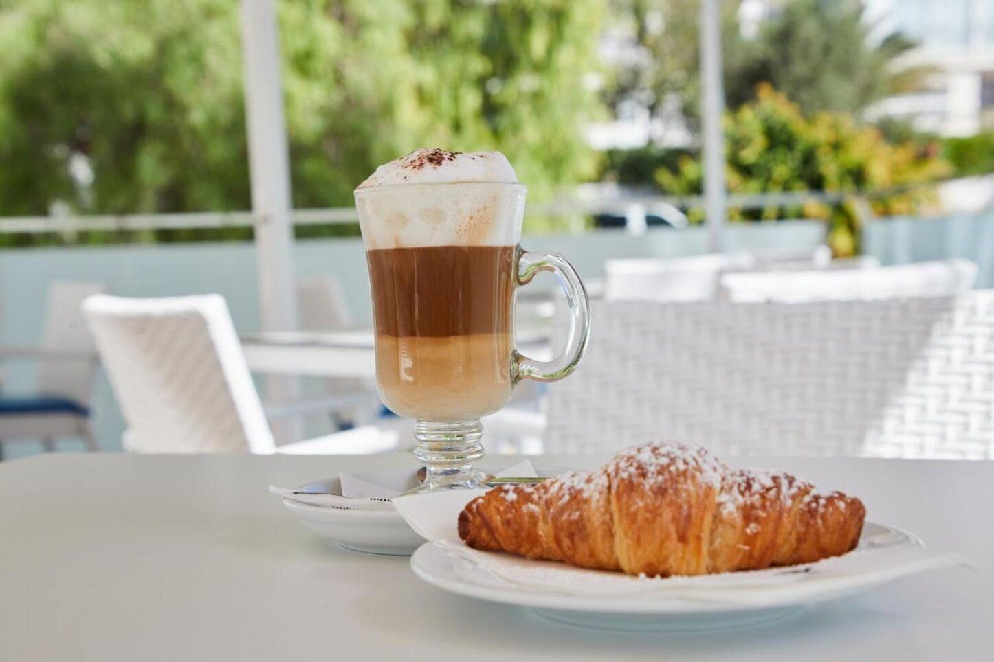 A glass of layered beverage with a pastry on a plate, set on a table with a white chair and a view of greenery.