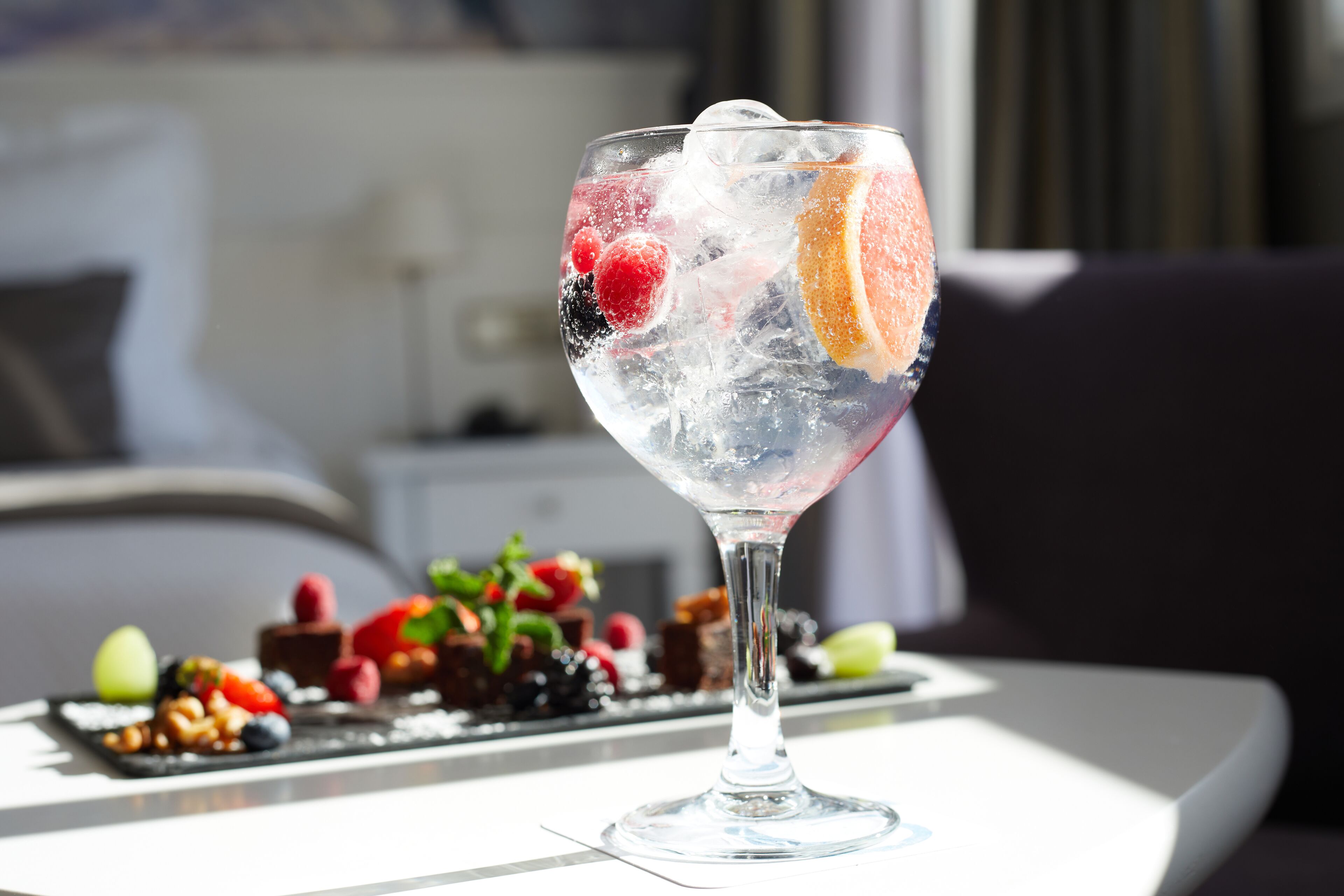 Un verre transparent rempli de glace et de fruits, une assiette de dessert avec des baies et du chocolat, et une pièce moderne avec une table blanche et un canapé foncé.