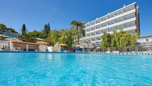 Una piscina con sillones de playa y sombrillas, rodeada por un edificio con varios balcones y árboles.