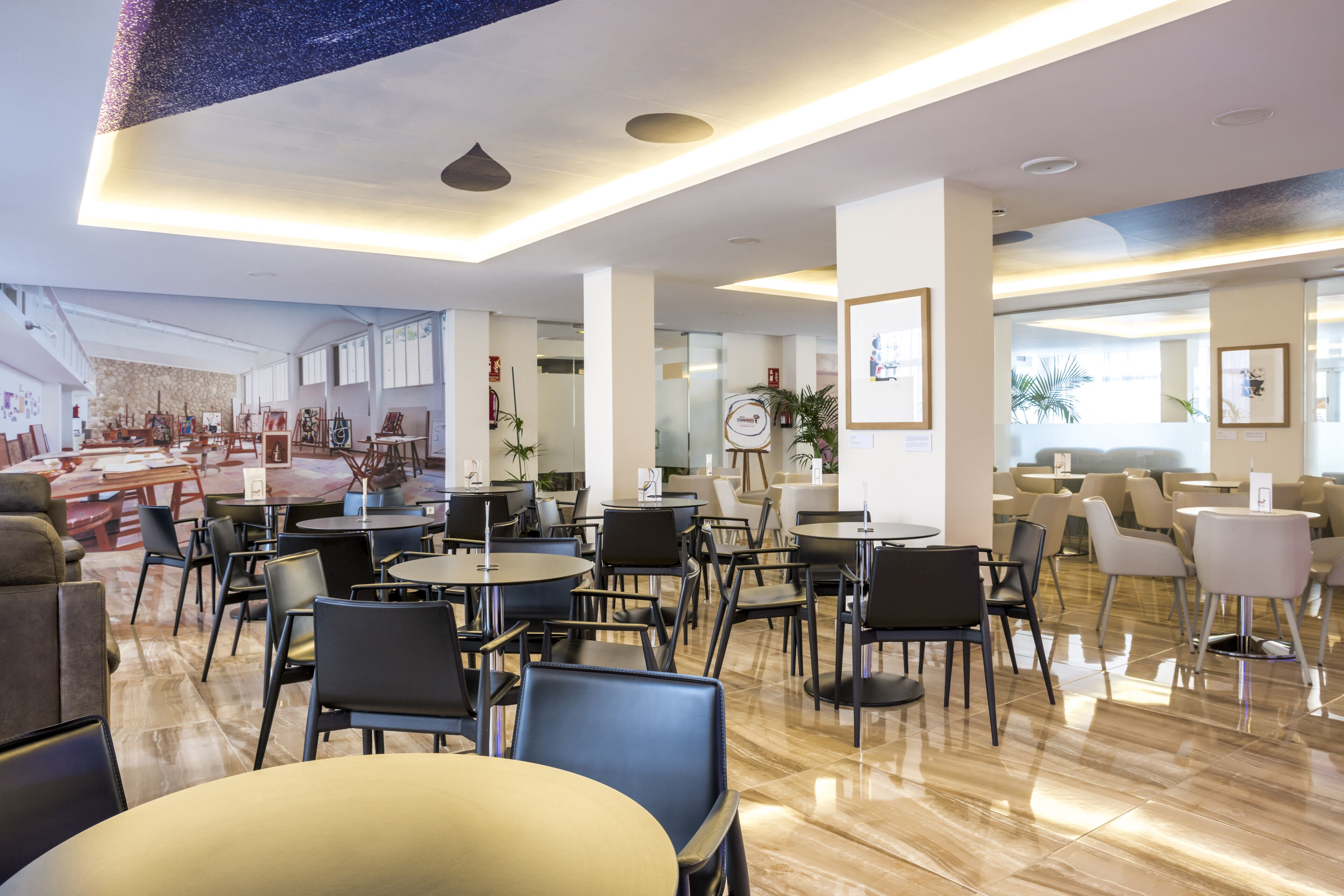 Un comedor moderno con mesas y sillas redondas, un techo alto con iluminación empotrada y vista del comedor al fondo.