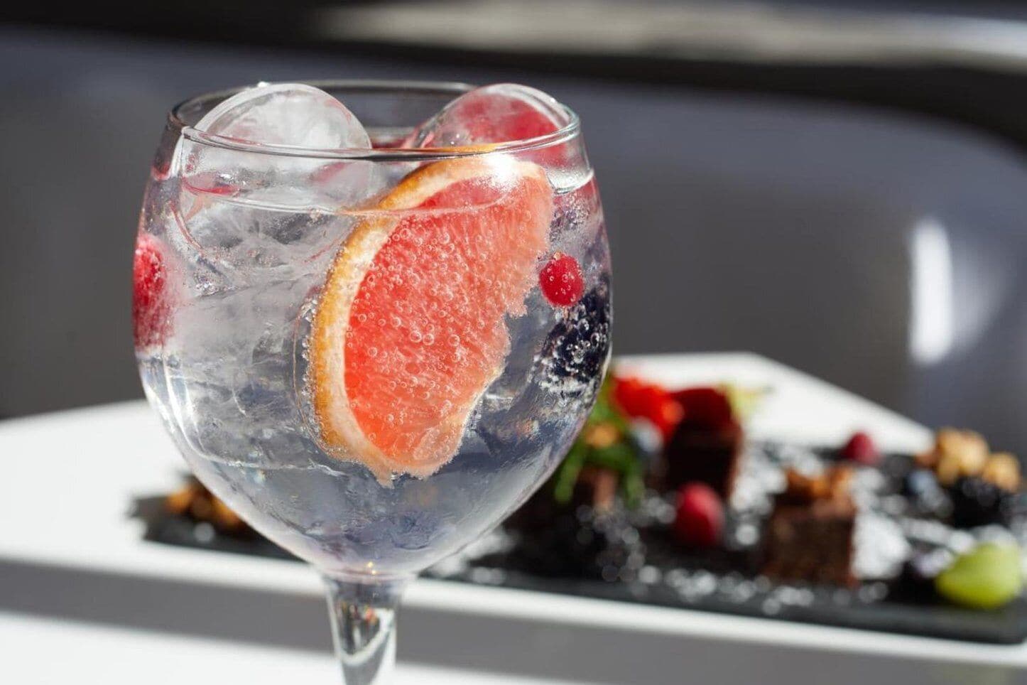 Un verre rempli de glaçons et une tranche de pamplemousse, avec une assiette de nourriture floue en arrière-plan.