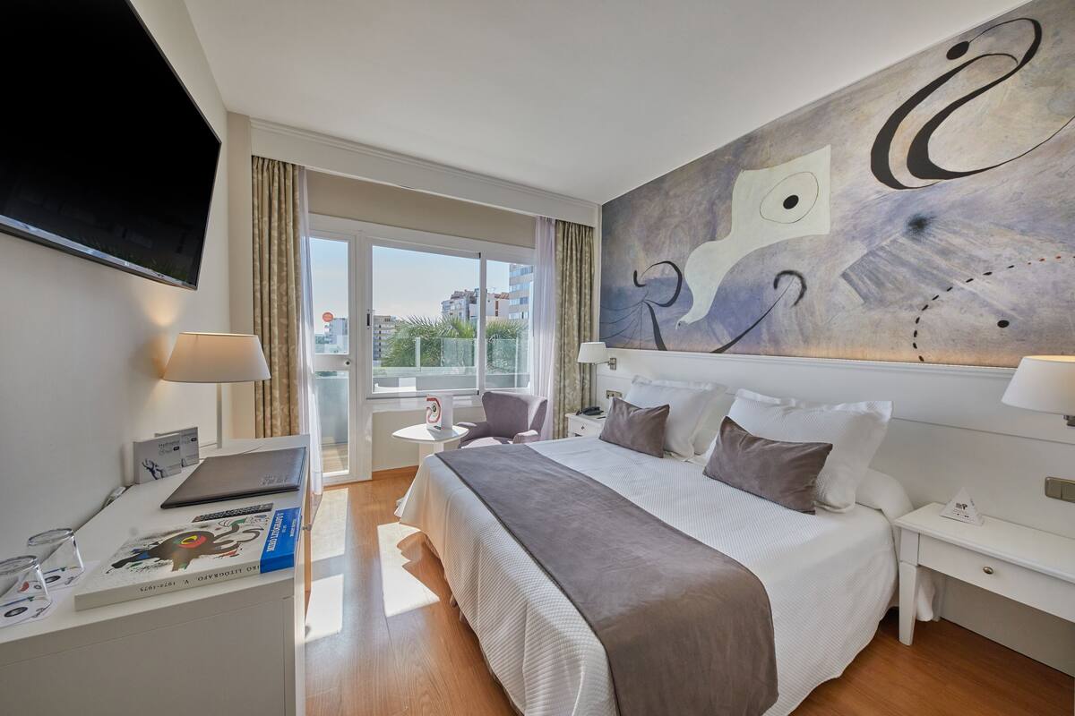 Un hotel moderno con una cama grande, un escritorio con una computadora portátil, un televisor de pantalla plana y vista a la ciudad por la ventana.