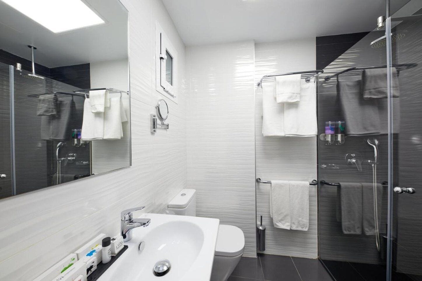 Un baño moderno con un espejo grande, baldosas blancas, un lavabo, inodoro y zona de ducha con mampara de vidrio.