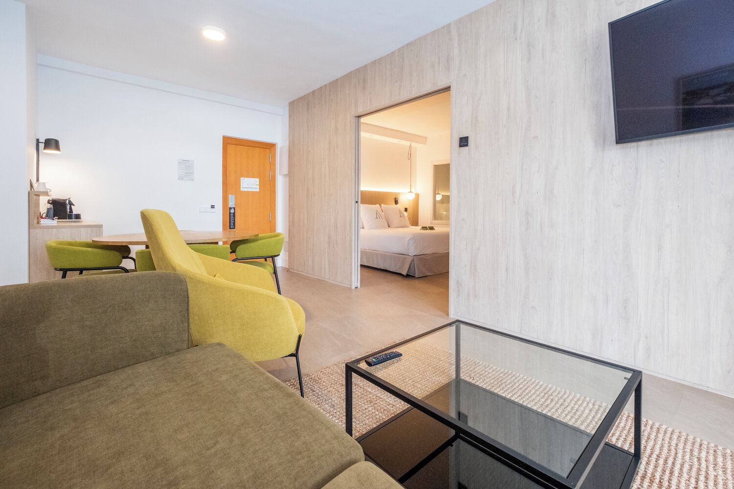Een moderne woonkamer met een bank, fauteuils, een glazen salontafel en een aan de muur bevestigde televisie.