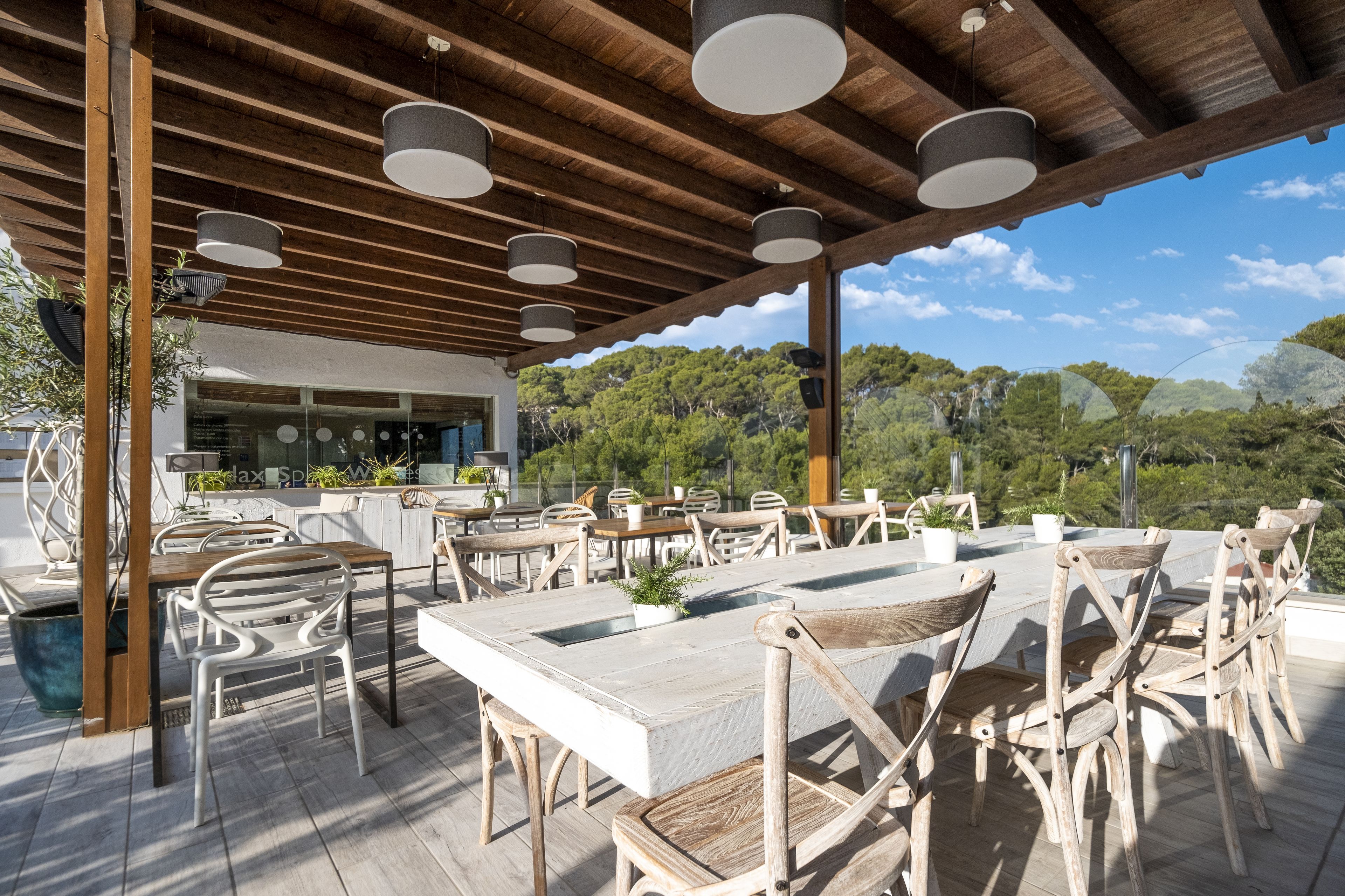 Un área de comedor al aire libre con mesas y sillas de madera, una pérgola con luces colgantes y vistas a una ladera boscosa.