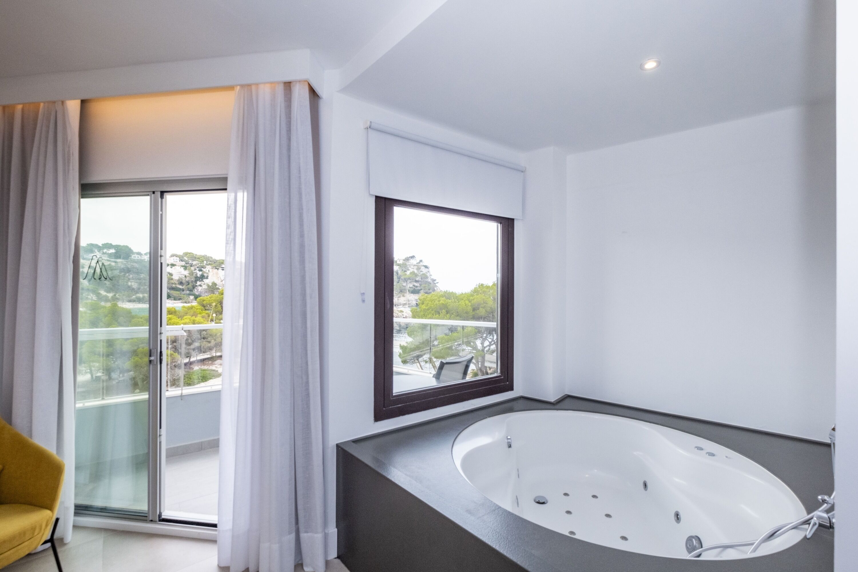 A modern bathroom with a freestanding bathtub, a large window with a view of the city, and a yellow chair.