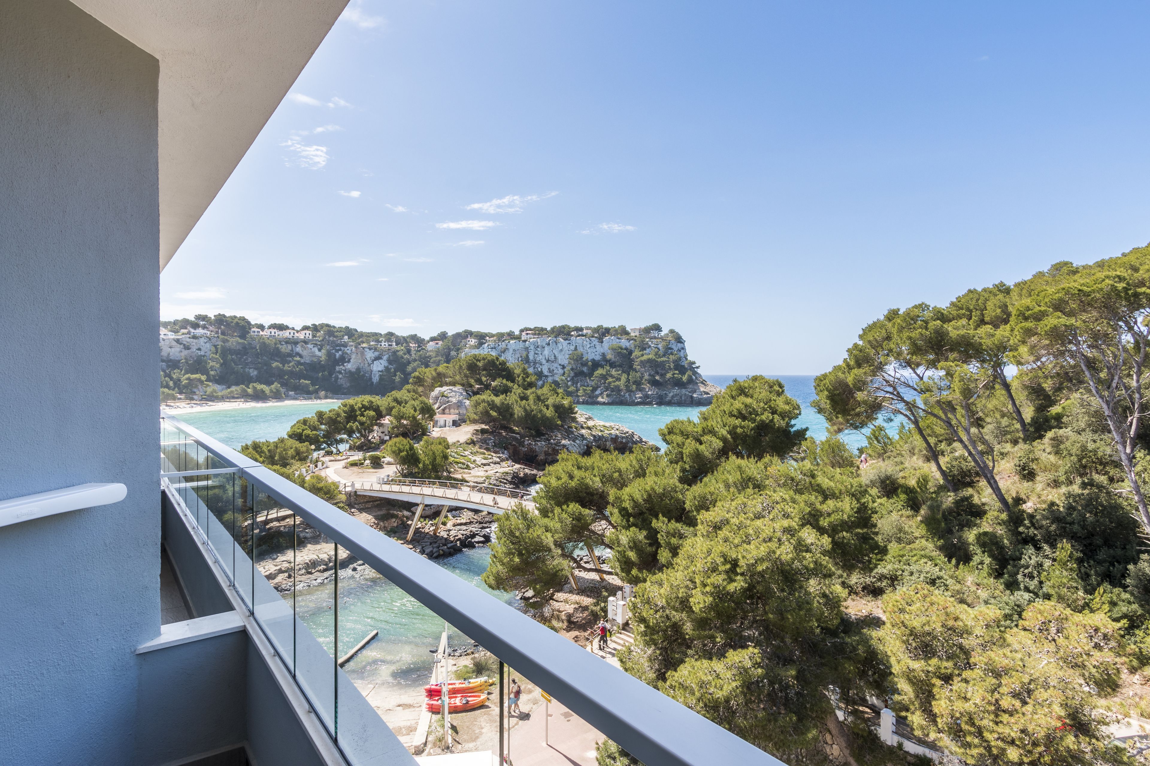 A balcony with a clear view of a coastal landscape, including a rocky beach, a bridge, and a cliff with buildings.