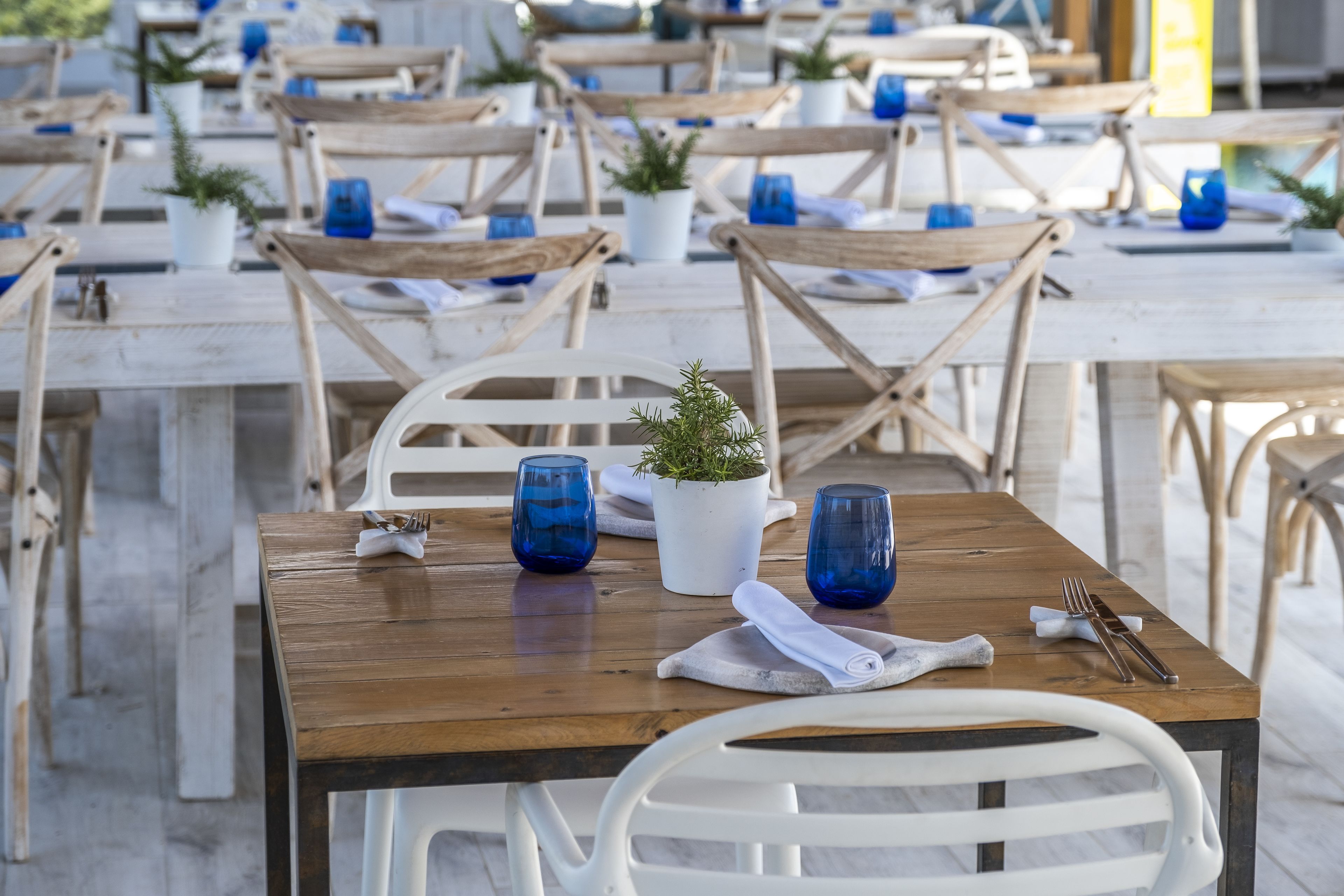 Un espace repas avec des tables en bois et des chaises blanches, certaines agrémentées de plantes en pot et décorées de verres bleus.