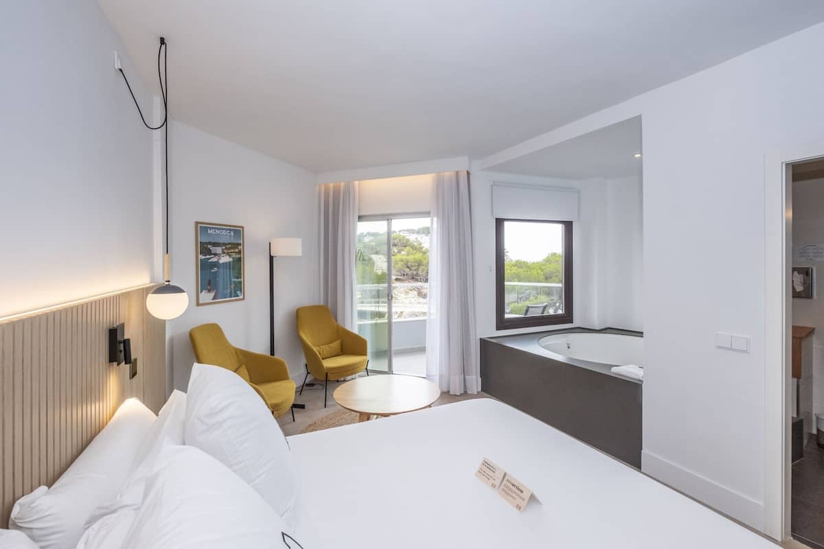 Une chambre d’hôtel avec un lit, une table ronde, un fauteuil jaune, une grande fenêtre et une salle de bain avec une baignoire.