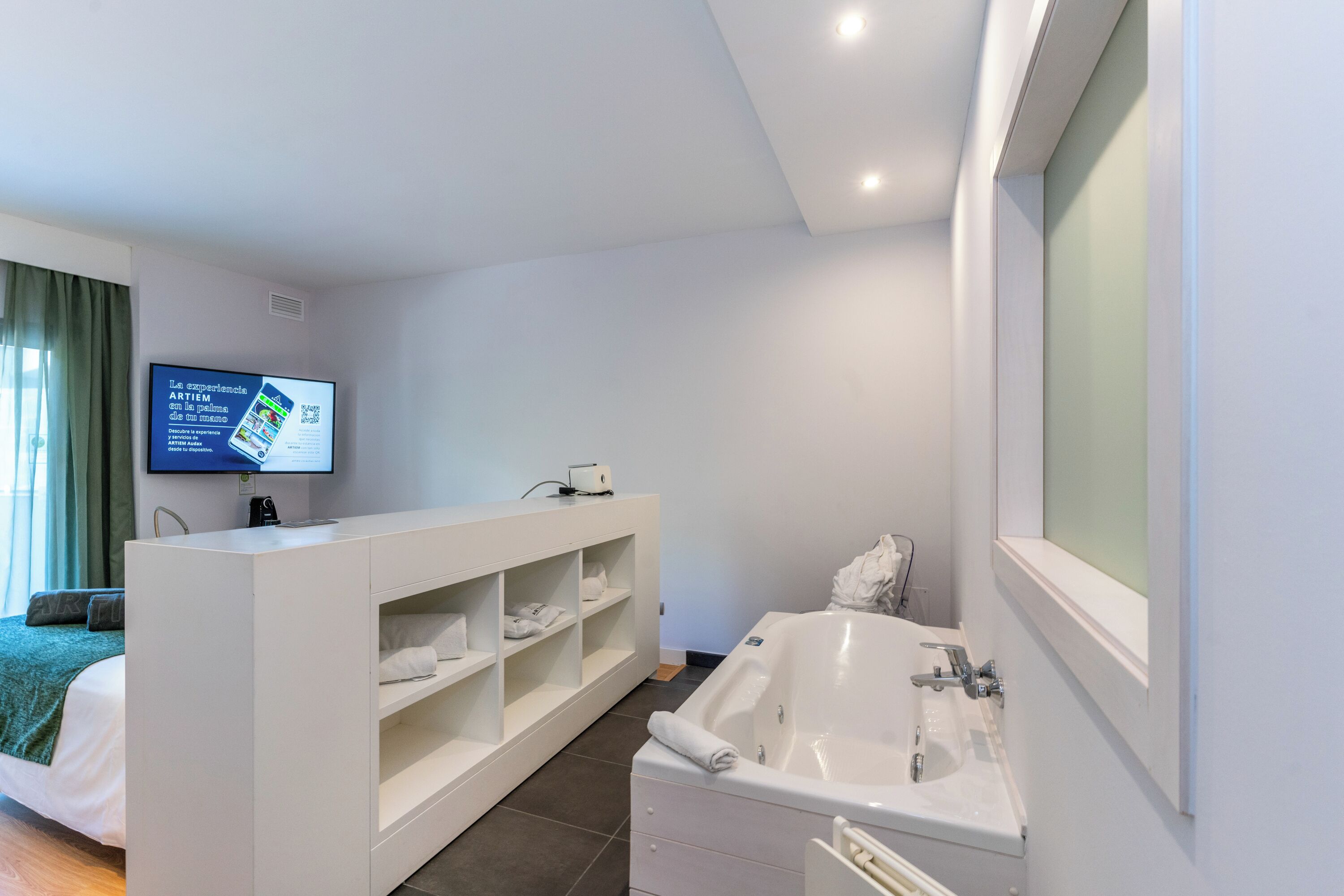 A modern hotel bathroom with a large bathtub, a vanity with shelves, and a flat-screen TV mounted on the wall.