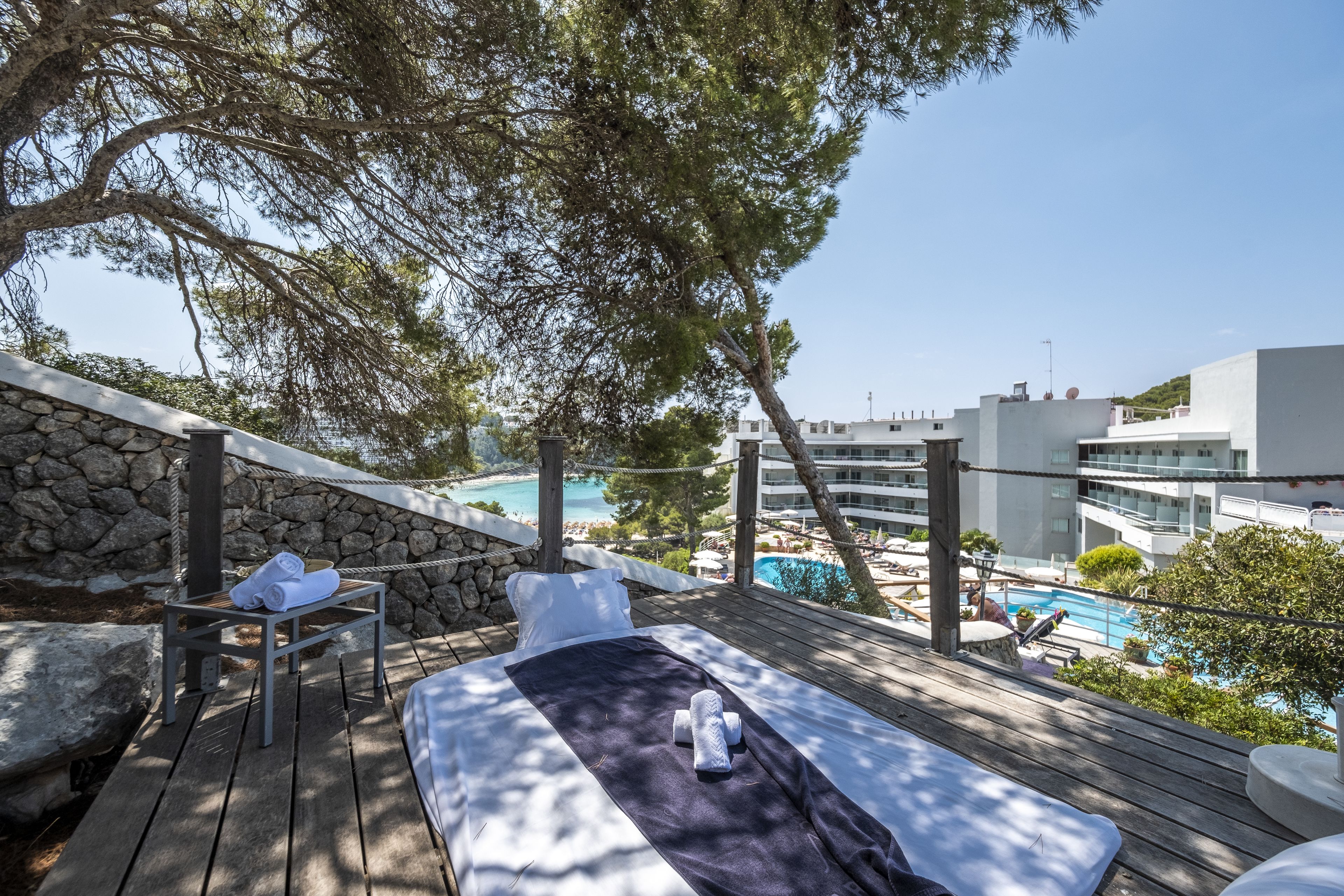 A wooden deck with a bed, a stone wall, and a view of a pool and buildings.