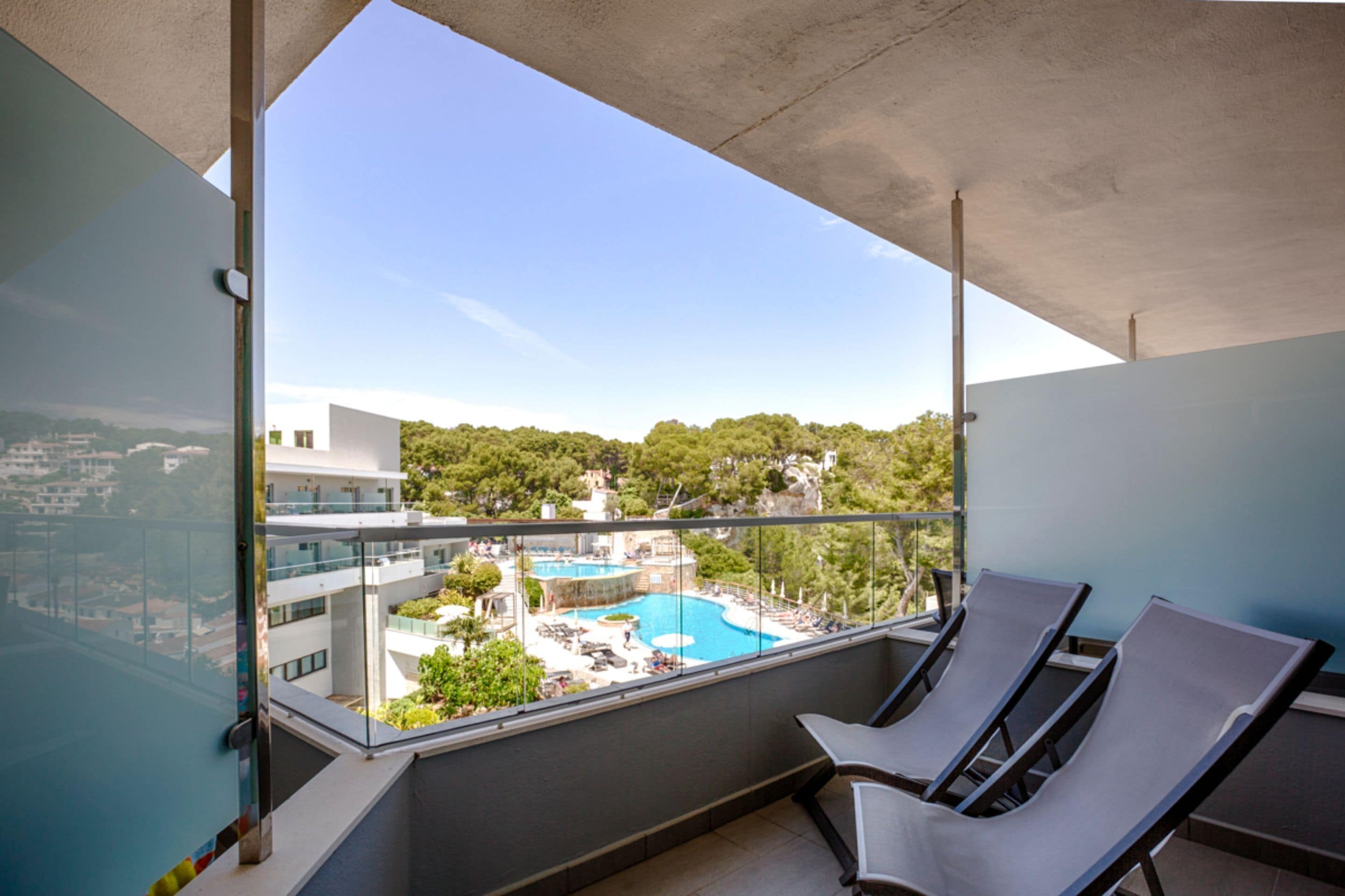 Un balcón con dos sillones de descanso, vista despejada a una piscina y un paisaje verde exuberante al fondo.