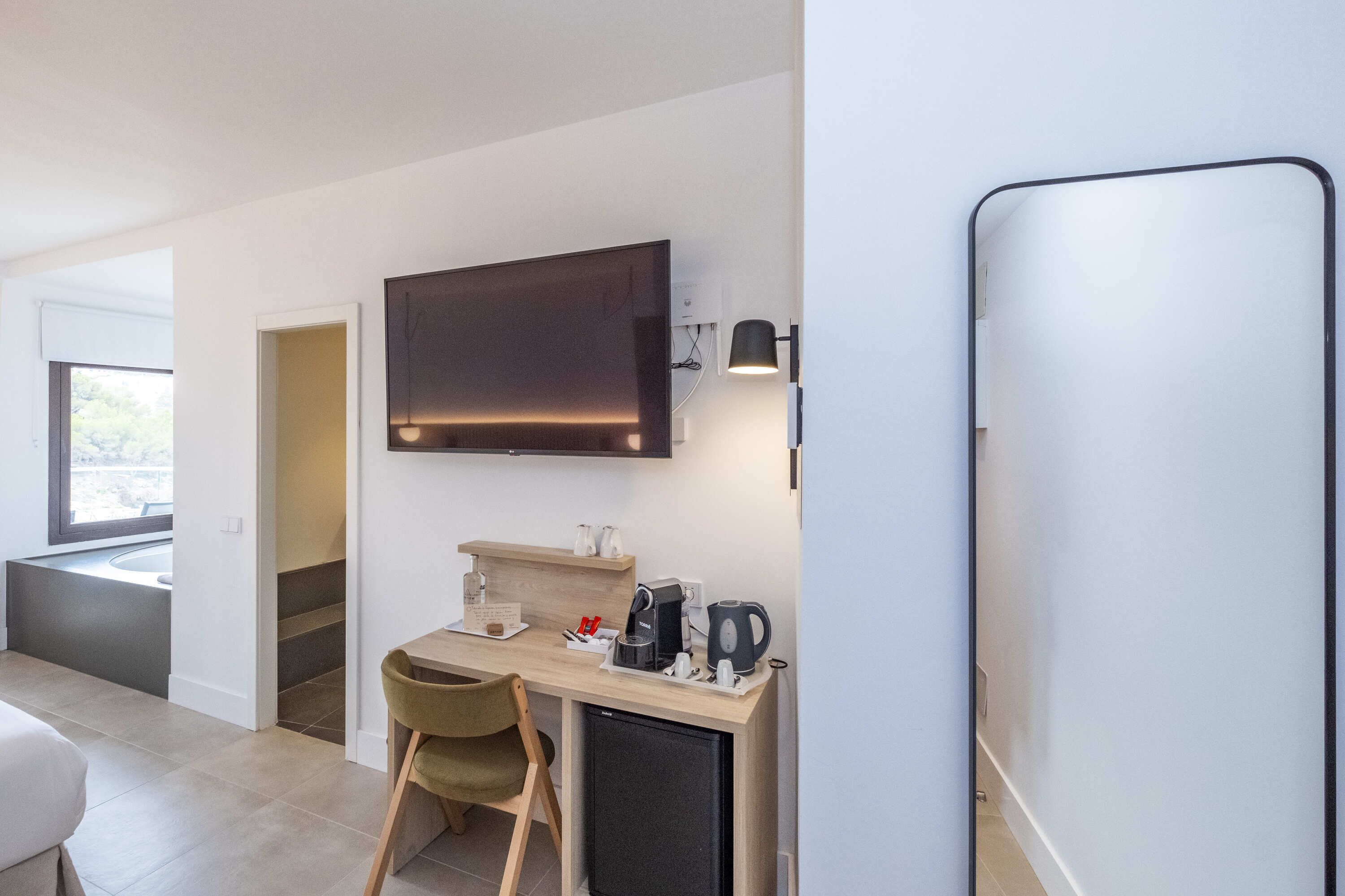 Una habitación de hotel moderna con una televisión de pantalla plana sobre un escritorio de madera, un refrigerador pequeño y una zona de estar con una silla.
