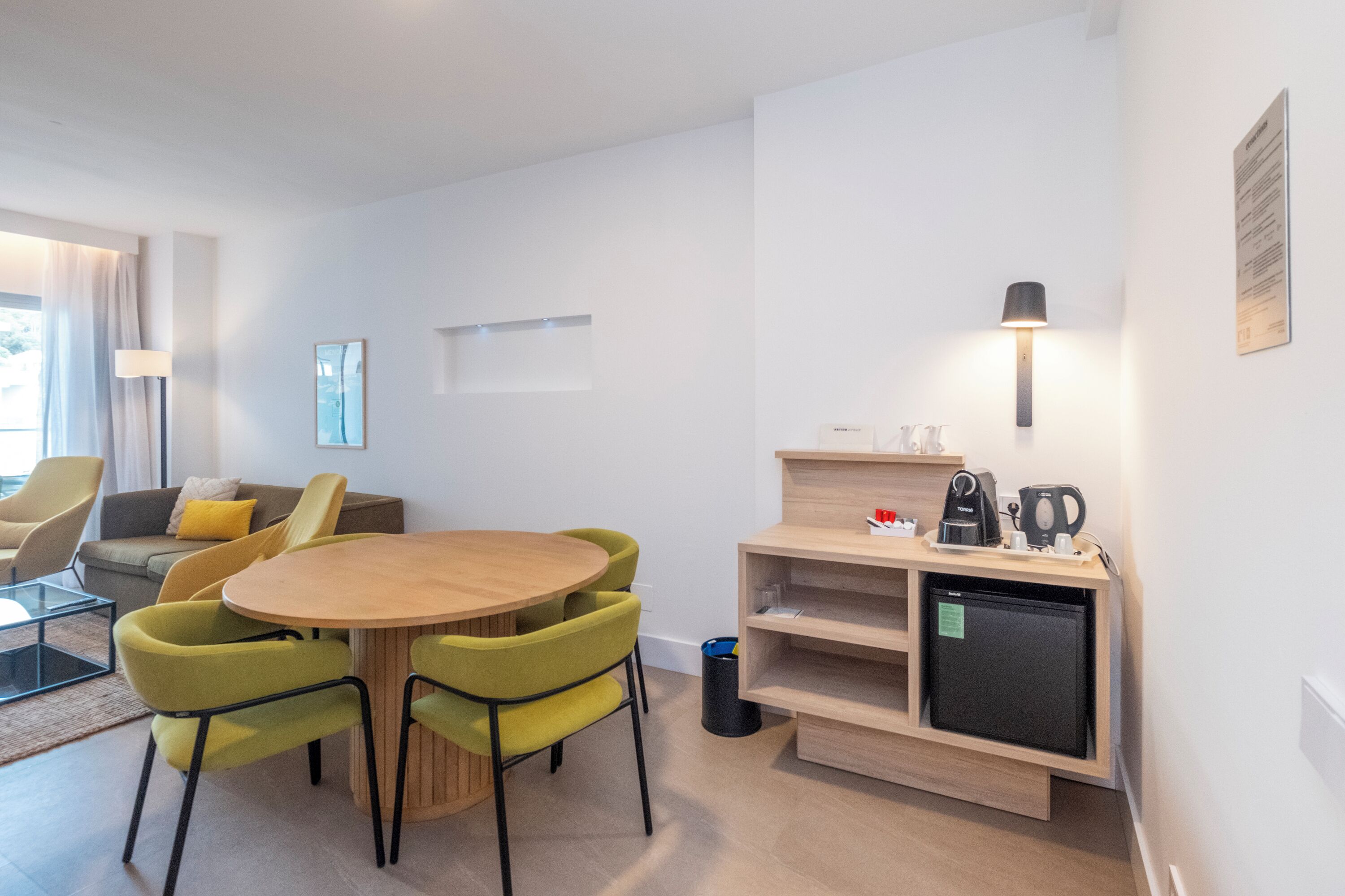 Una habitación moderna y luminosa con una mesa redonda de madera rodeada de sillas verdes, un refrigerador negro pequeño y un mueble estantería empotrado con una lámpara.