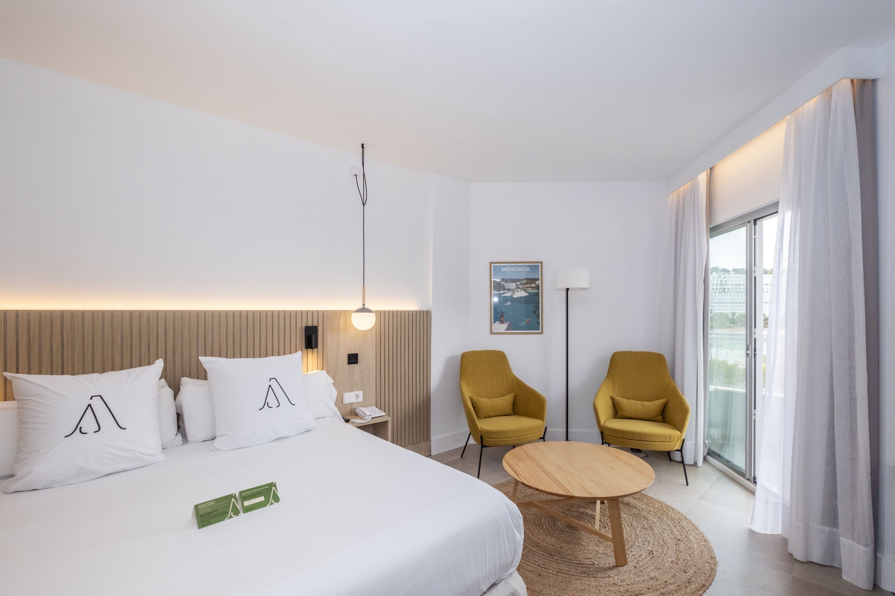 Un hotel moderno con una cama grande, dos sillones amarillos, una mesita de madera y vistas a la ciudad a través de un ventanal amplio.