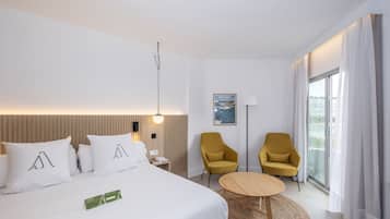 Un hotel moderno con una cama grande, dos sillones amarillos, una mesita de madera y vistas a la ciudad a través de un ventanal amplio.