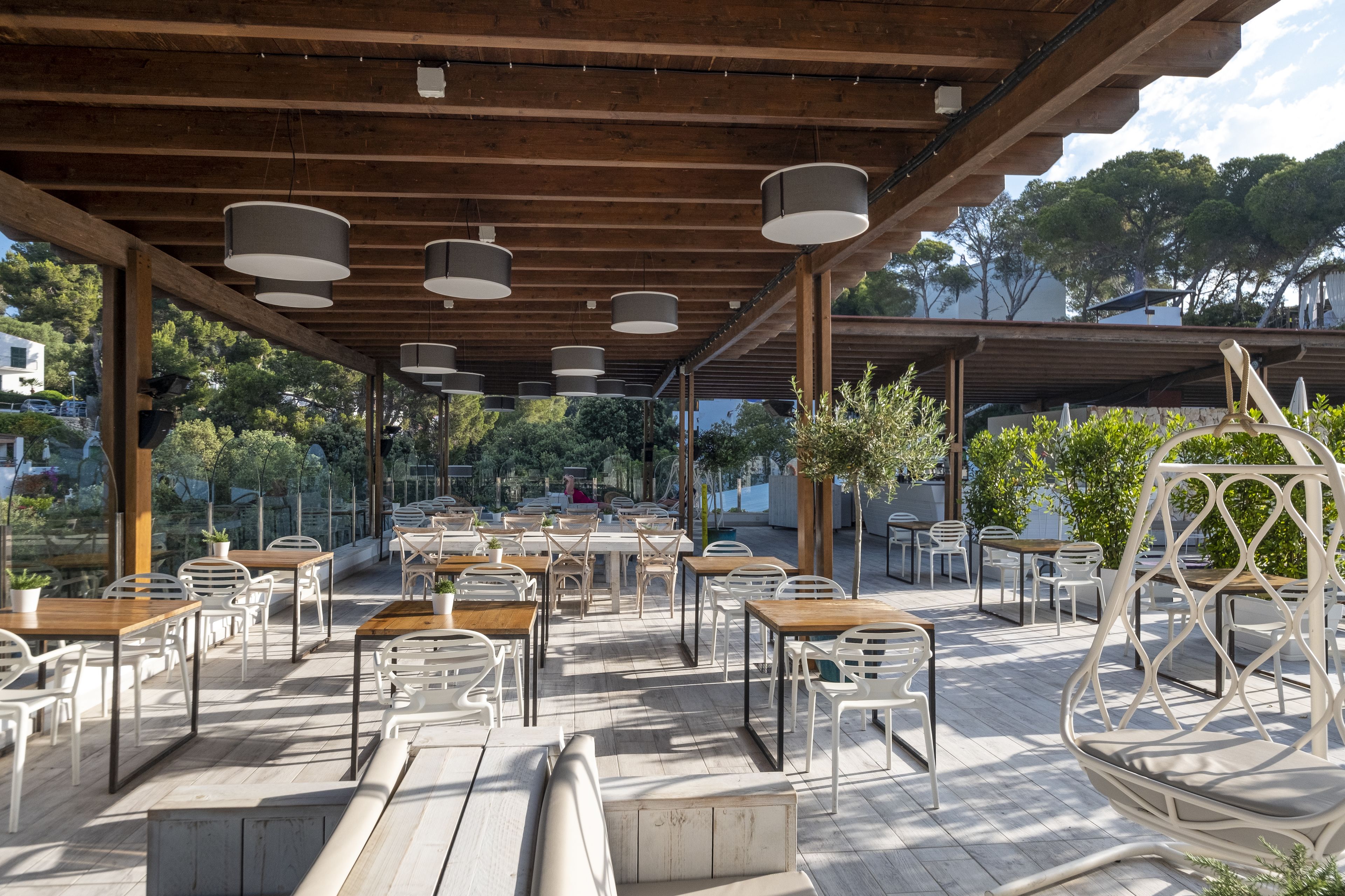 An outdoor patio with wooden tables and chairs, a covered seating area, and a view of trees and buildings in the background.