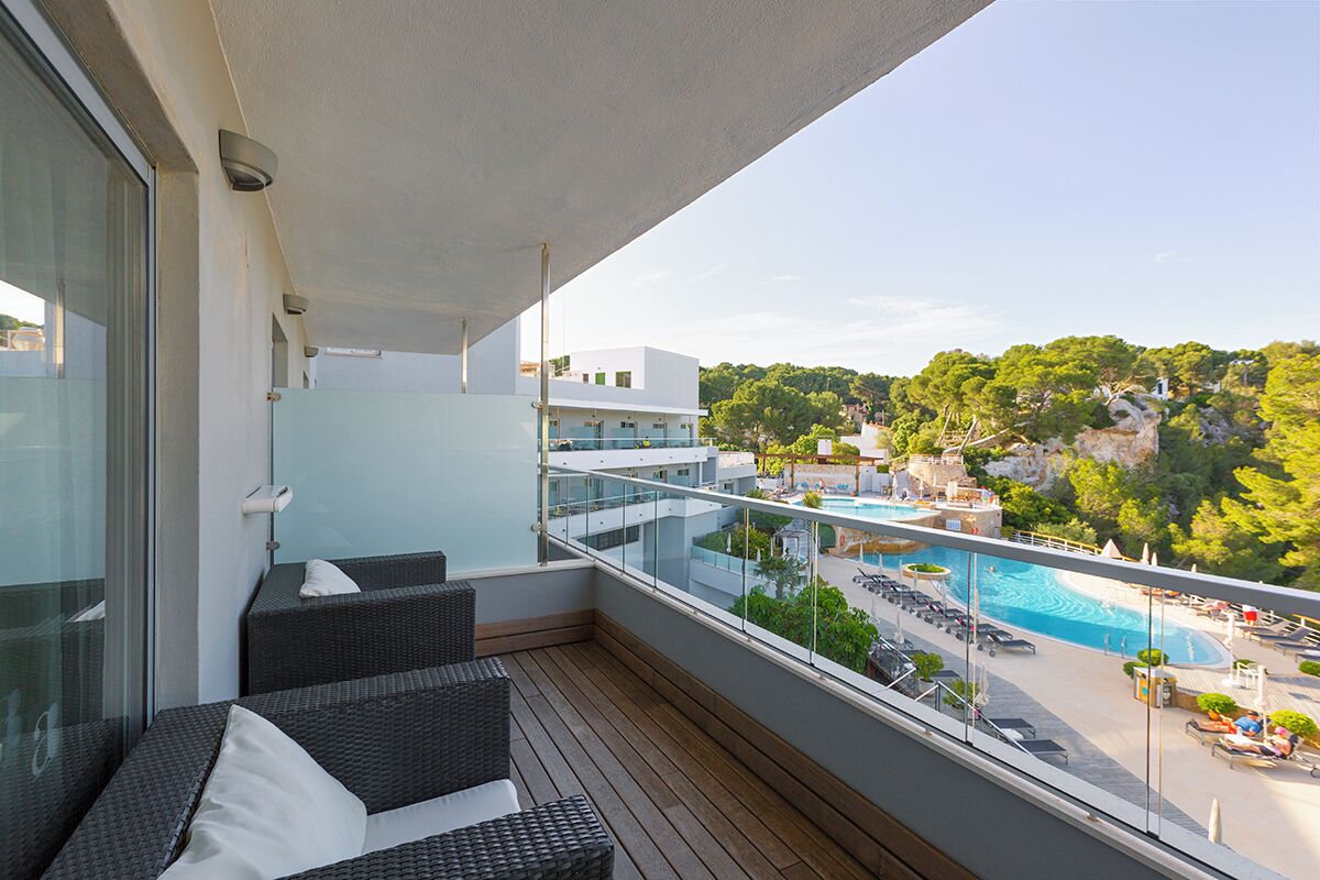 Ein Balkon mit Korbmöbeln, einer Glasreling und Blick auf einen Pool und Grünflächen.