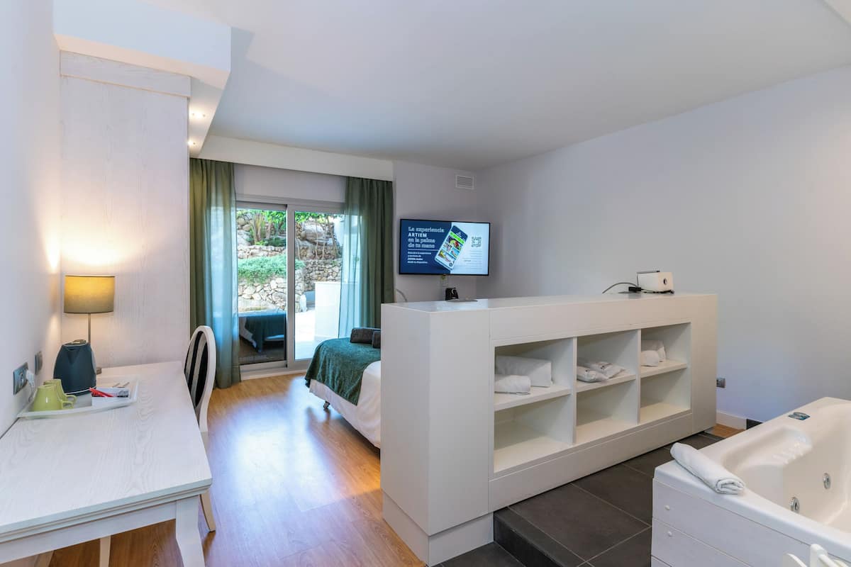 Une chambre d’hôtel moderne équipée d’un lit, d’un bureau, d’une télévision, d’une baignoire et offrant une vue sur la verdure environnante.