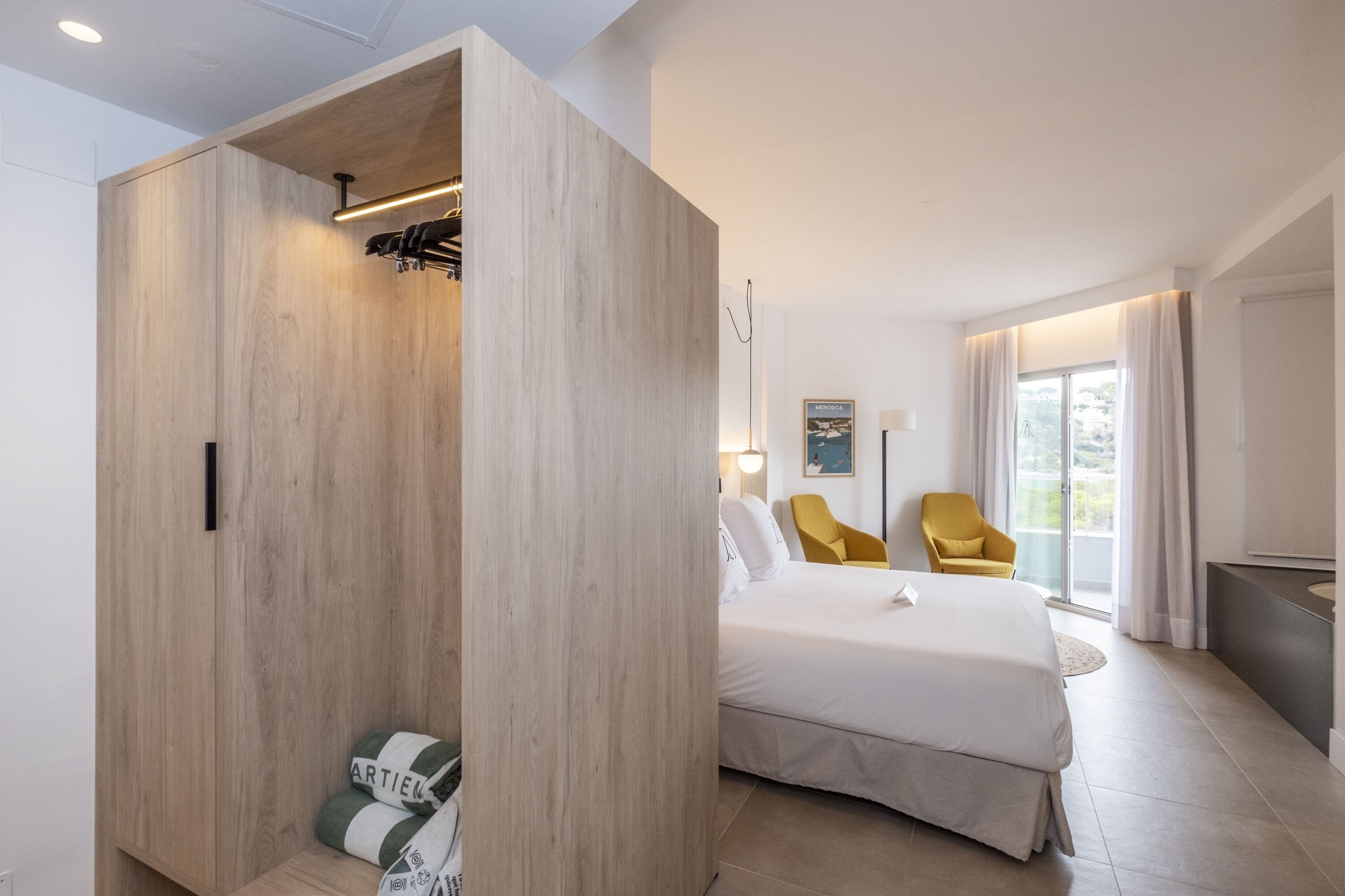 Habitación de hotel moderna con un amplio armario de madera, una cama con ropa blanca, un sillón amarillo y un ventanal con cortinas.
