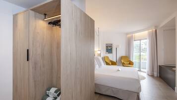 Habitación de hotel moderna con un amplio armario de madera, una cama con ropa blanca, un sillón amarillo y un ventanal con cortinas.