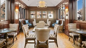 A dining area with a central round table surrounded by upholstered chairs, wooden paneling, and large windows offering views of buildings. - Poste Montane Lodge (Avon)
