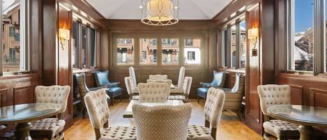 A dining area with a central round table surrounded by upholstered chairs, wooden paneling, and large windows offering views of buildings.