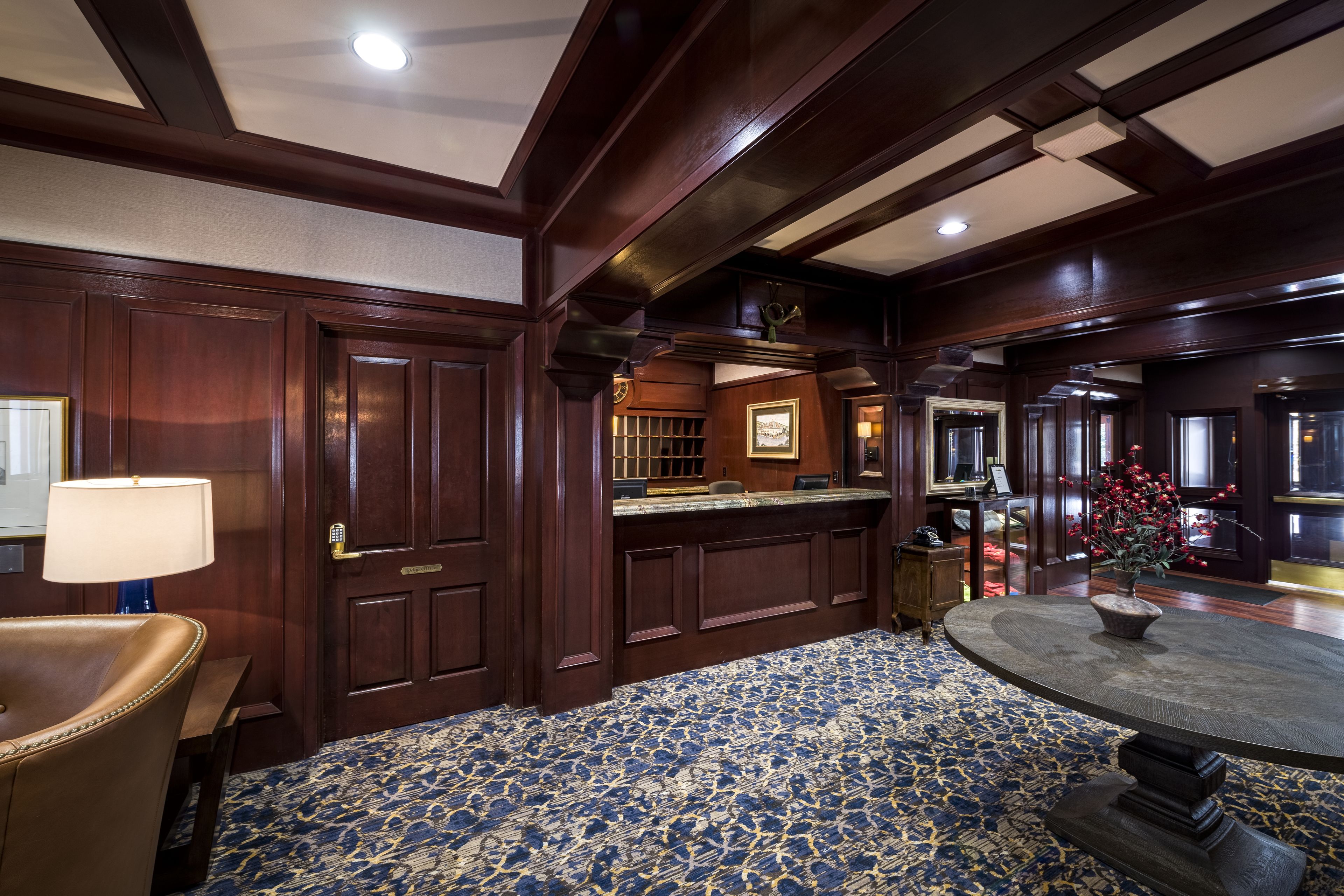 A hotel lobby with dark wood paneling, a round table with a vase, a blue patterned carpet, and a desk with a lamp.