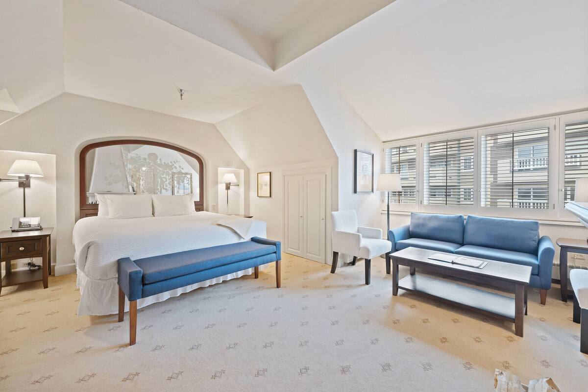 A hotel room with a large bed, a blue bench, a white armchair, a blue sofa, and a small table.