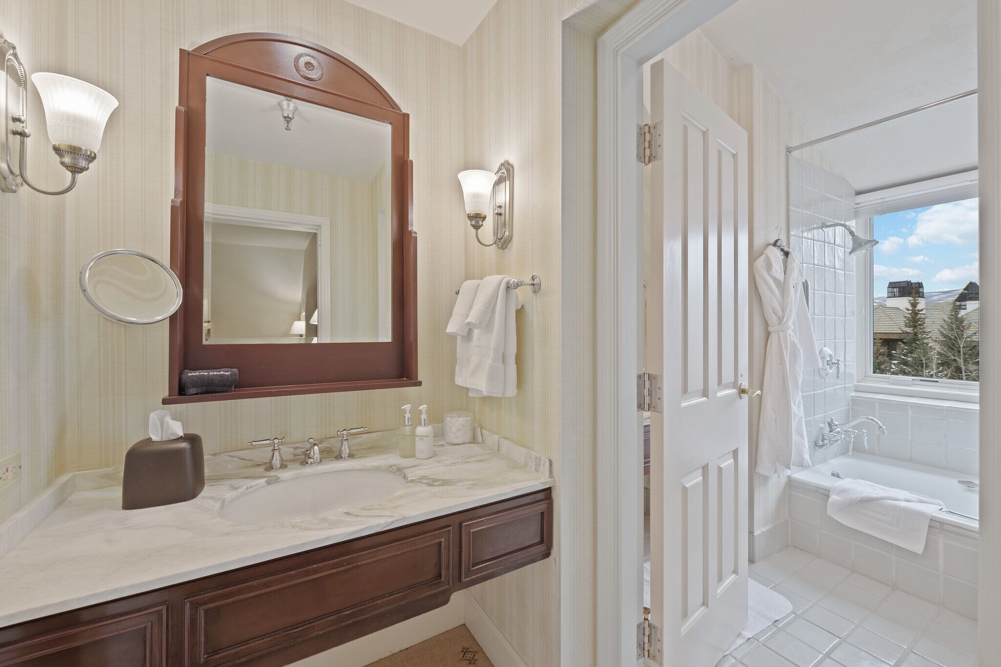 A bathroom with a double sink, a large mirror, a towel rack, and a view of the outside through a window.