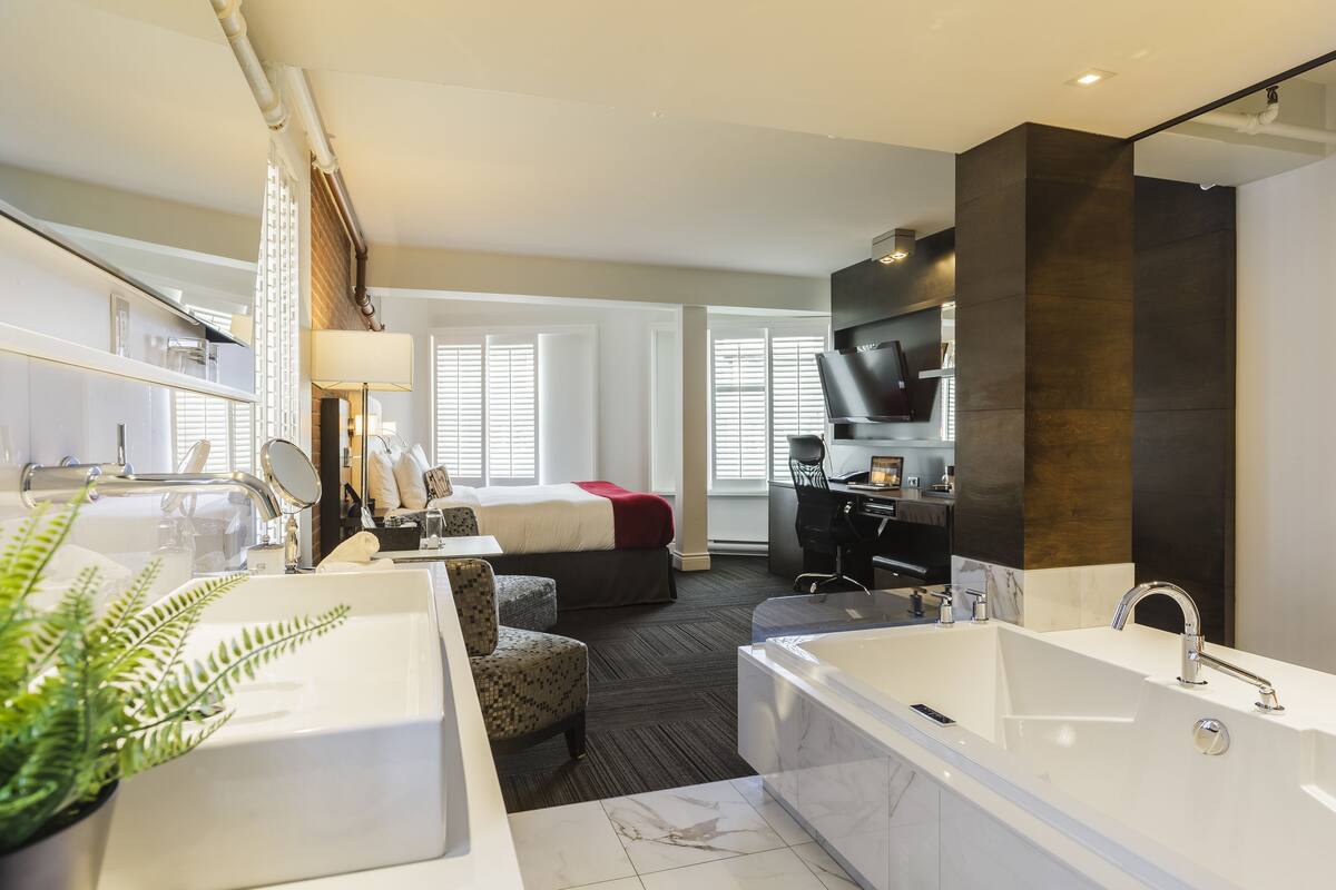 A modern hotel room with a large bathroom featuring a bathtub, a separate shower area, and a sink. There is a bed with a red blanket, a small seating area with a chair, and a TV mounted on the wall.