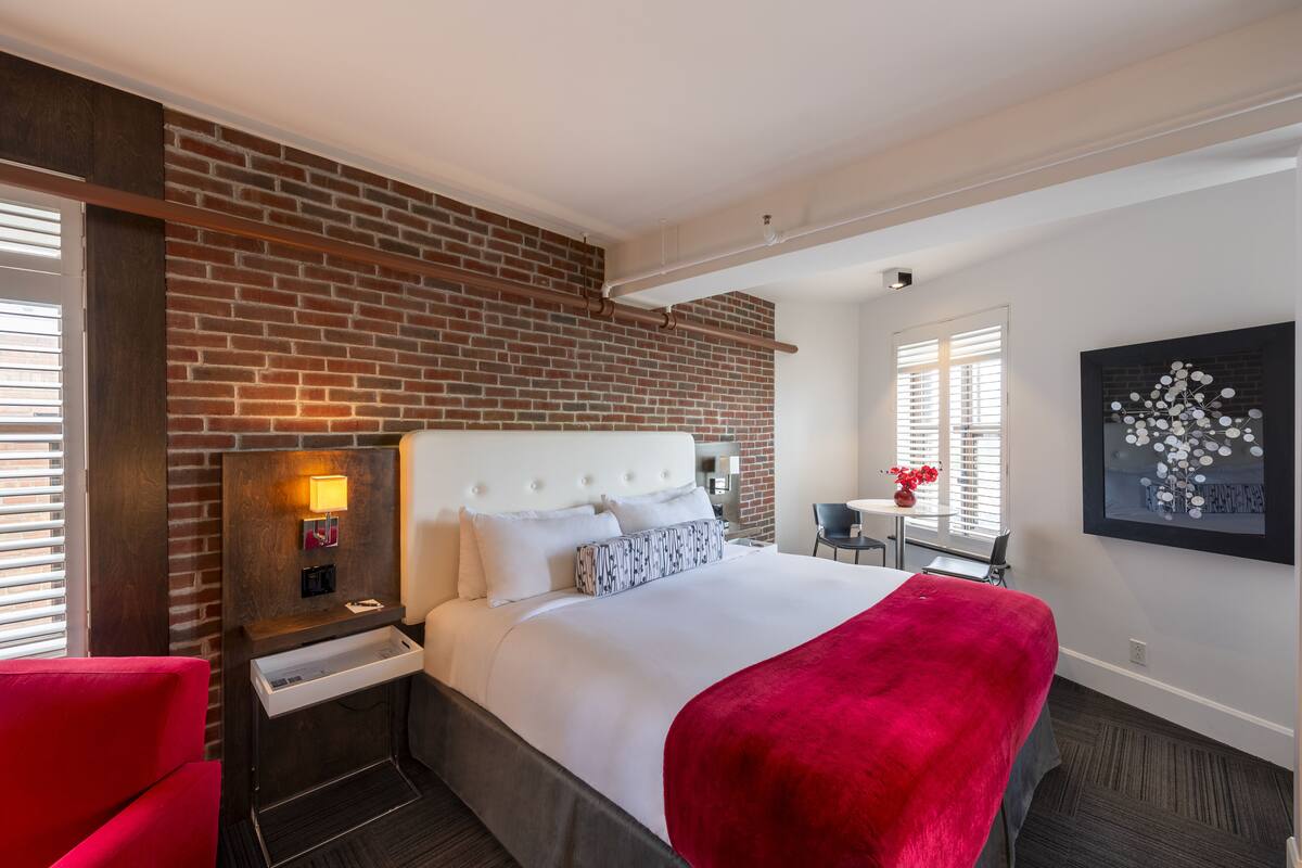 A hotel room with a brick accent wall, a white bed with a red throw, a nightstand with a lamp, and a small table with a vase.
