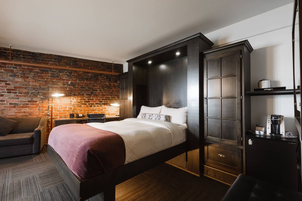 A modern hotel room with a brick wall, a bed with a headboard, a wardrobe, a desk with a lamp, and a coffee machine.
