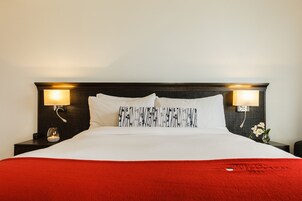 Una cama bien hecha con ropa de cama blanca y roja, dos lámparas de noche y un pequeño jarrón con flores en la mesita.
