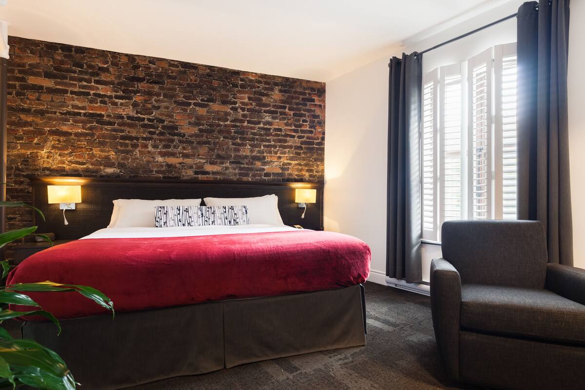 A hotel room with a brick wall, a large bed with a red blanket, two bedside lamps, a chair, and a window with curtains.