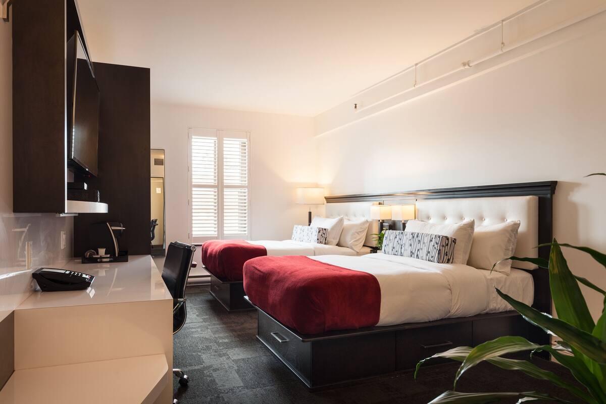 A hotel room with two beds, a desk, a TV, and a window with blinds.